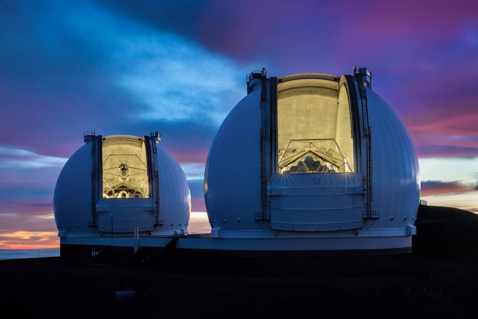 Image of light from two large telescopes pouring out onto a pink, sunset sky