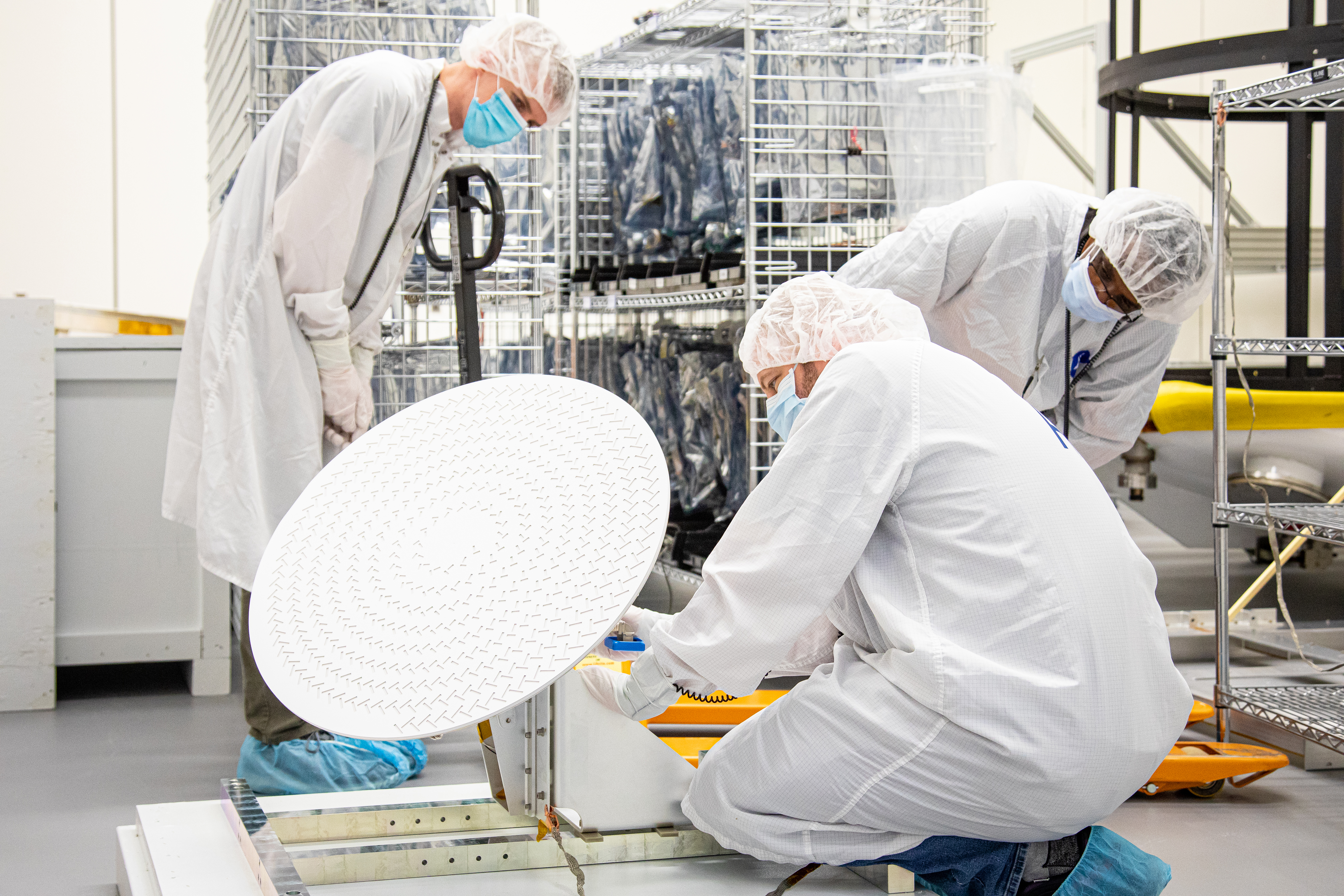 Three people in personal protective gear look over a piece of spacecraft