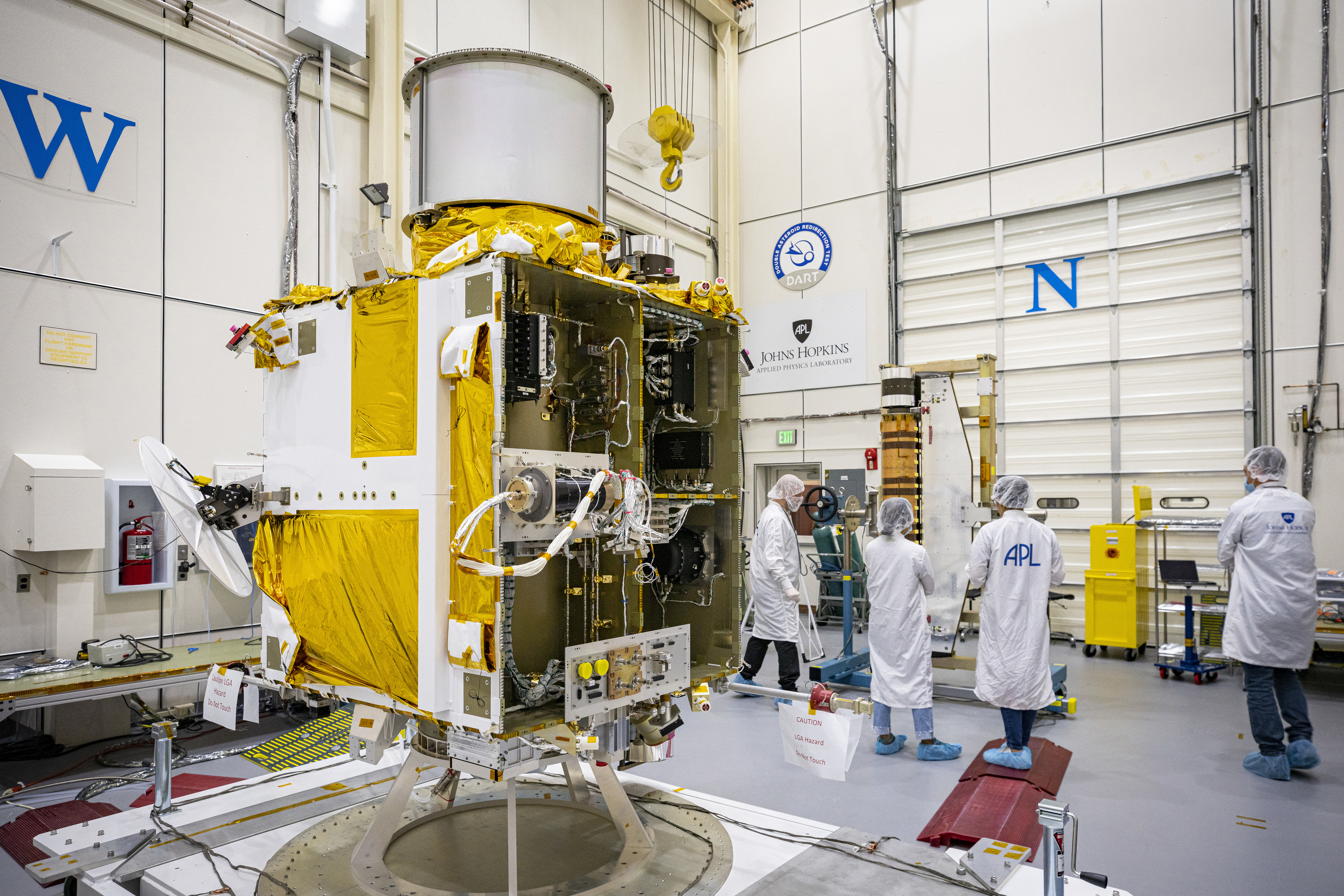 People in personal protective equipment stand next to the gold-foil-covered DART spacecraft