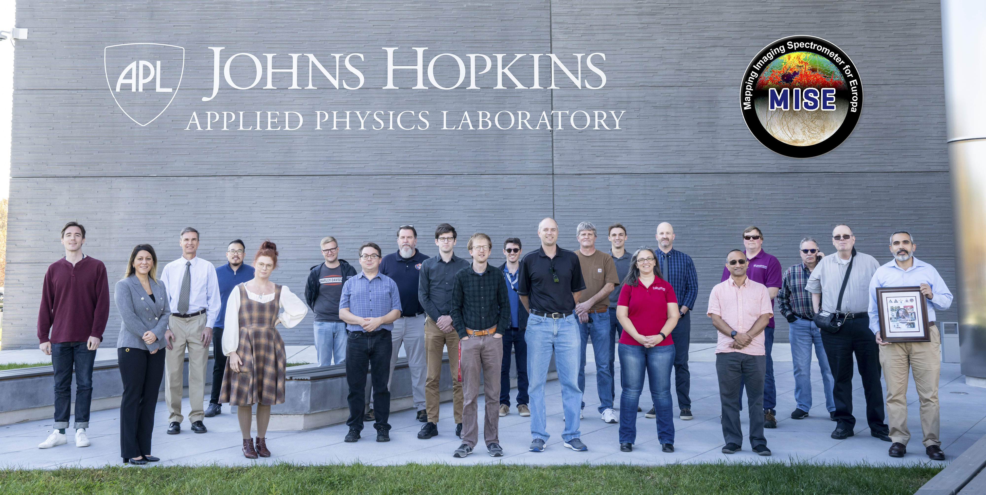 People stand outside of a building with Johns Hopkins Applied Physics Laboratory branded on its wall