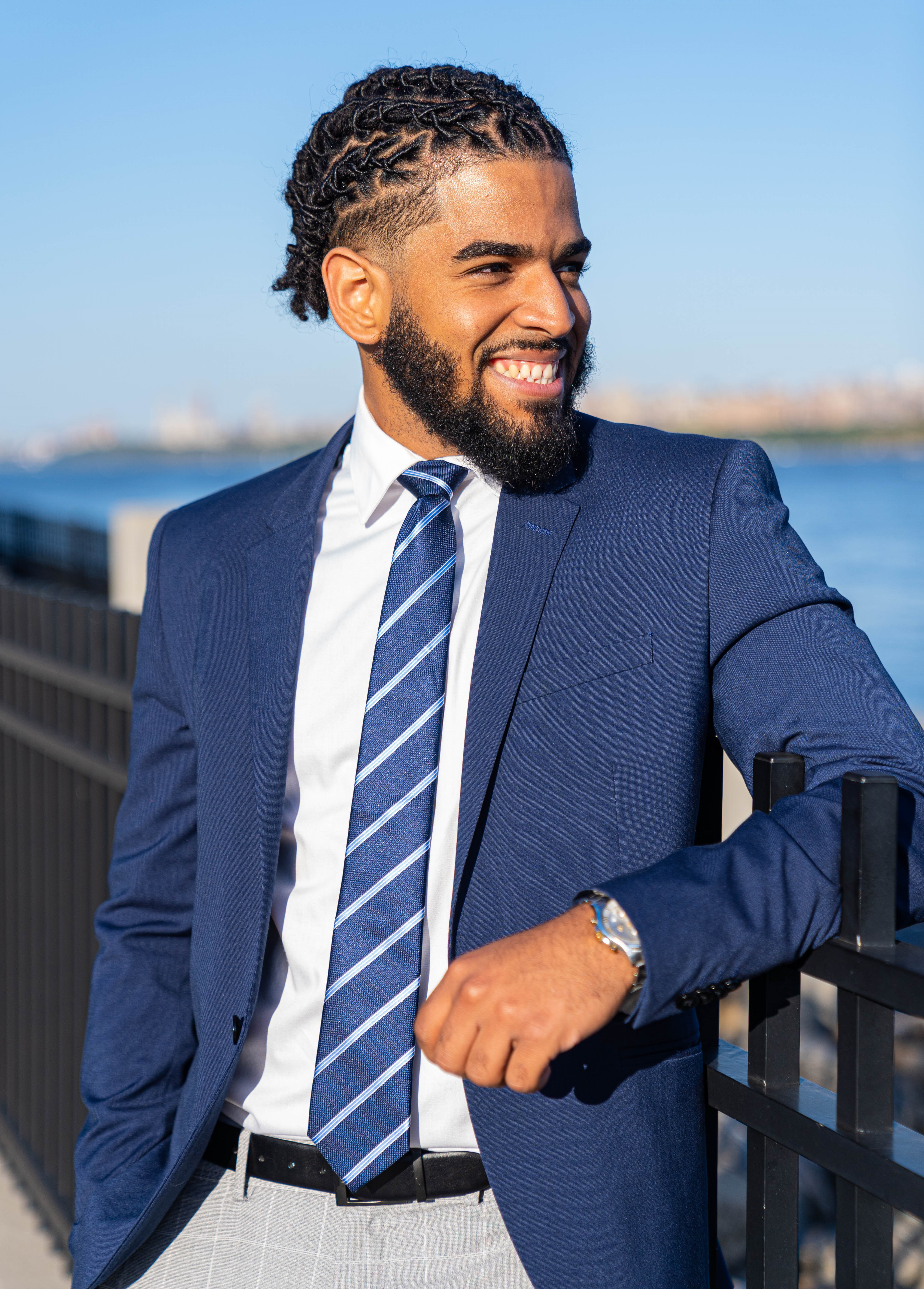 A Dominican man dressed in khakis, a blue blazer, white shirt and blue striped tie stands smiling while looking off to a body of water