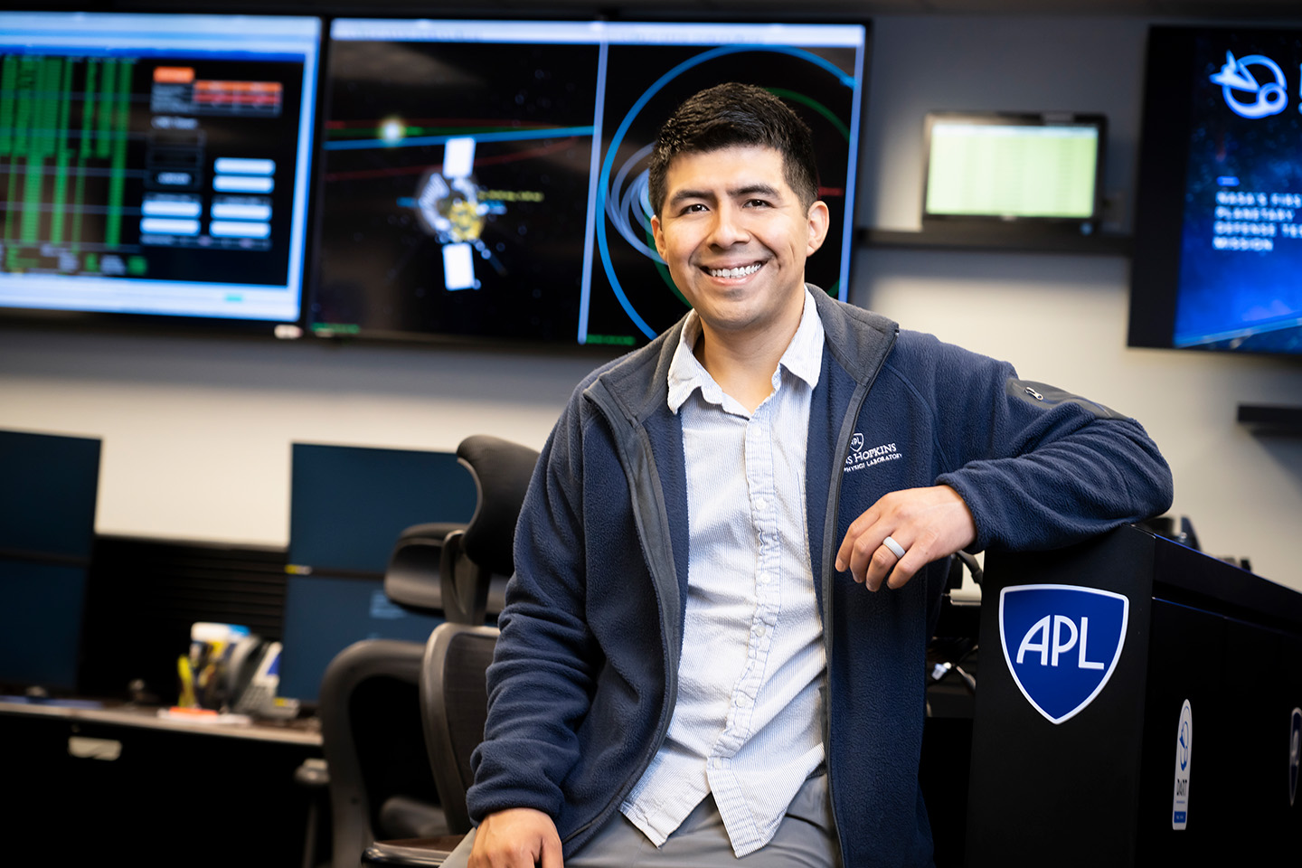 A man wearing a collared shirt and blue fleece jacket with the logo "Johns Hopkins Applied Physics Laboratory" on it sits in a room full of computers and TV screens, with stickers of the APL shield on some of them.