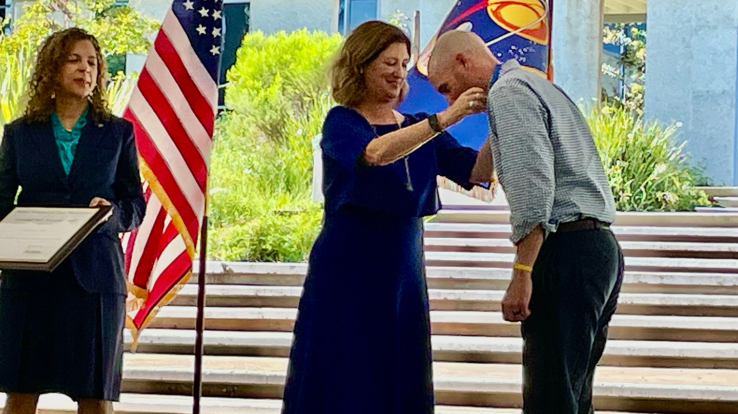 Outside, a man leans over as a woman in a blue dress places a medal around his neck. Another woman in a dress stands off to the side near a podium and microphone, with the United States flag behind her