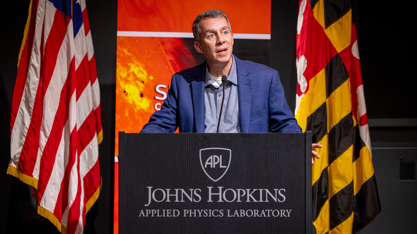 A photo of Bobby Braun standing at a podium between the American and Maryland flags, with a placard in front that says "Johns Hopkins Applied Physics Laboratory"