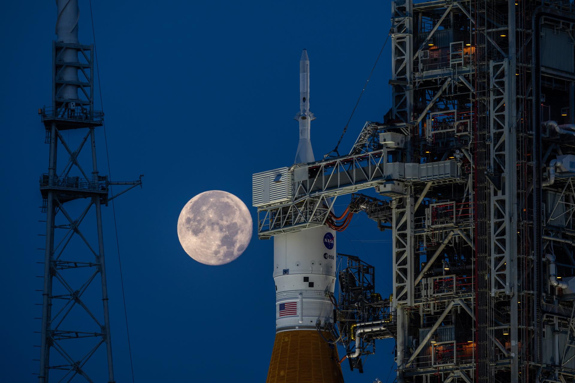 A photo shows the top of a rocket with a NASA symbol on it. Behind it is the full Moon