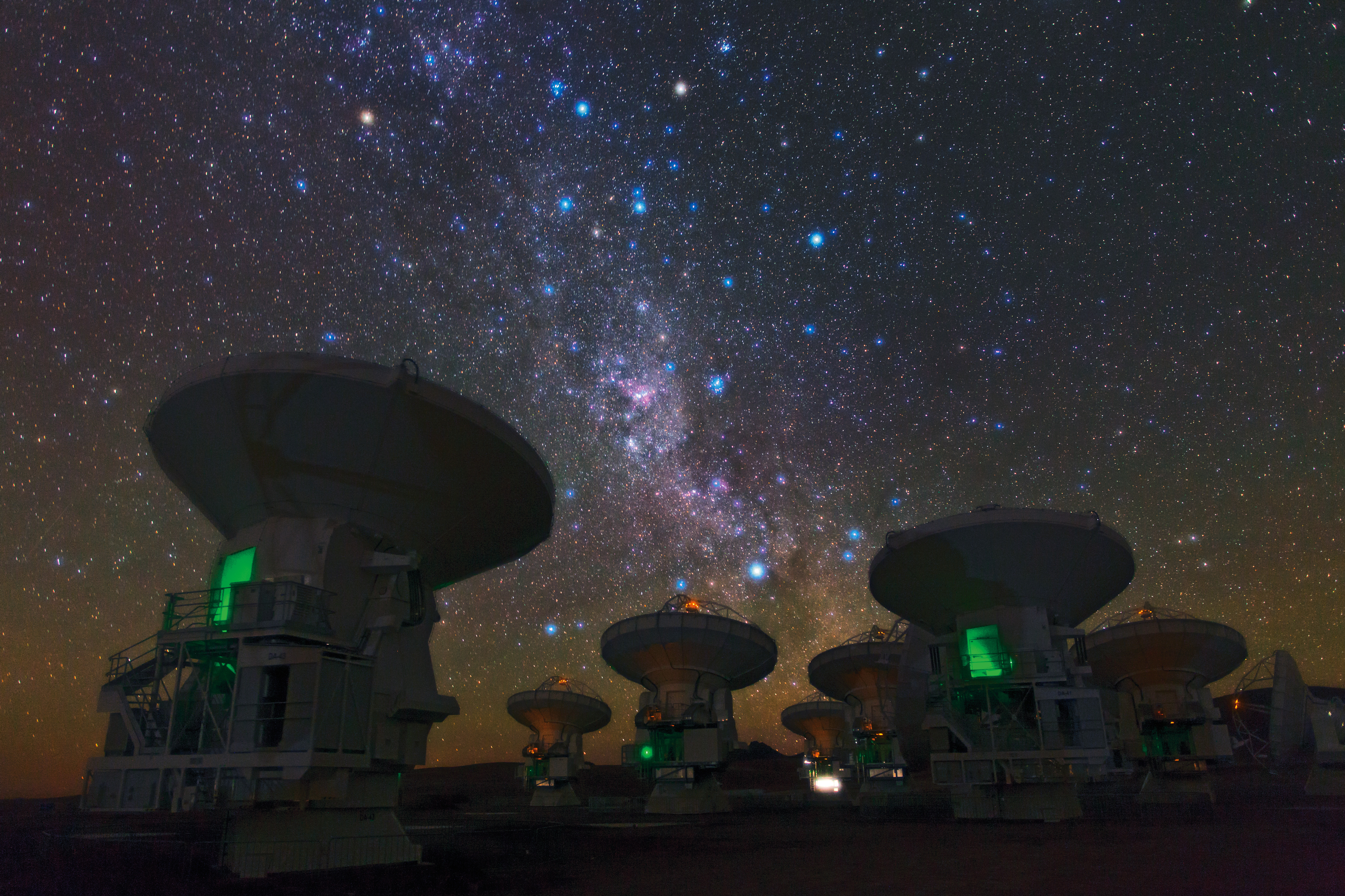 A nighttime photo shows satellite dishes on Earth pointing straight up into the starlit sky, where the Milky Way and various bright stars are conspicuously visible