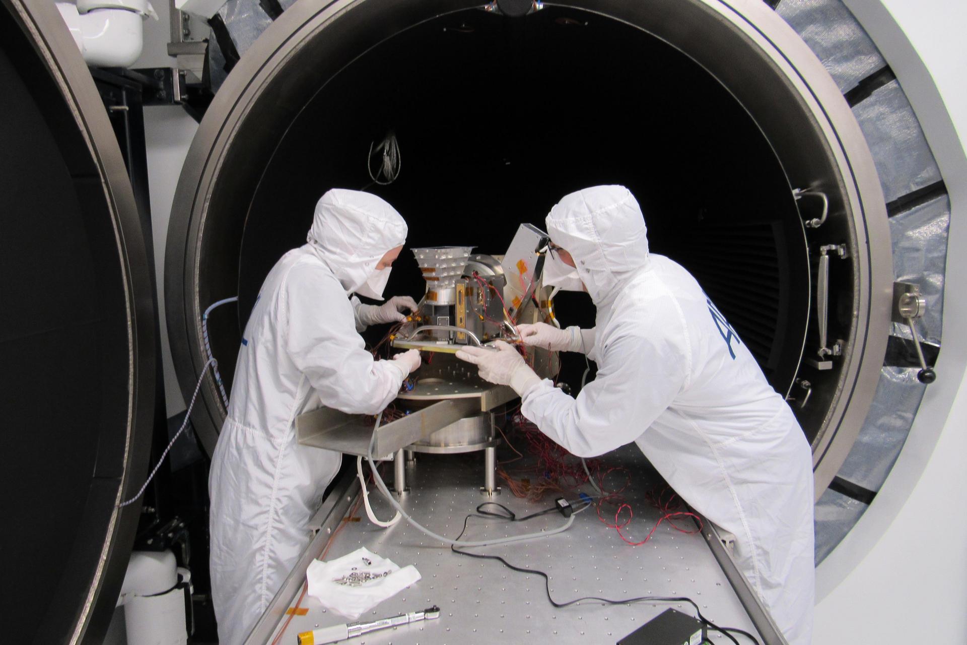 Two people in white clean suits work on an instrument 